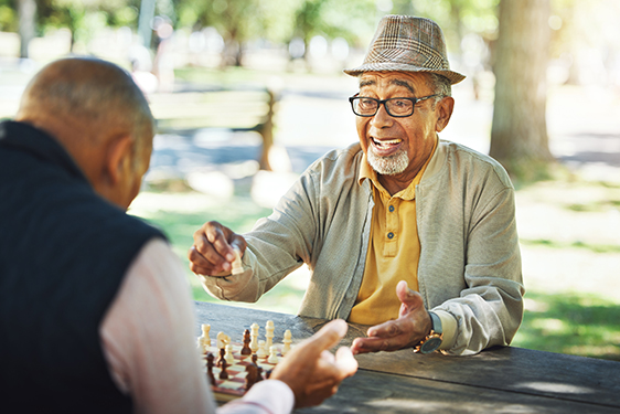 Two older Black men sit at an outdoor table and play chess. One is making a move and smiling, while we can only see the back of the other man. 
