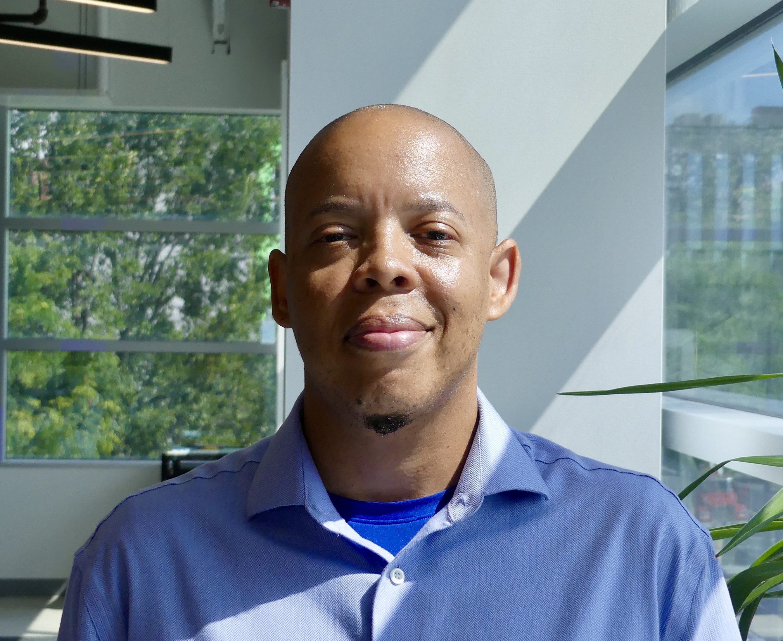 A Black person smiling at the camera wearing a blue collared shirt and lots of greenery in the background